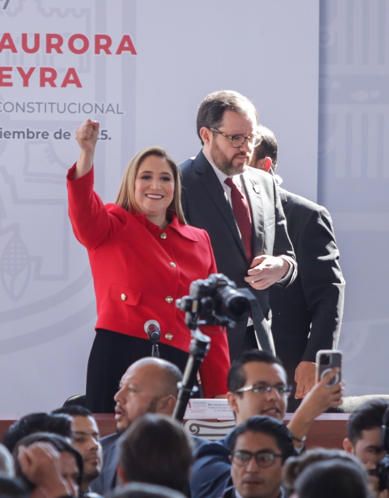 La presidenta municipal de San Mateo Atenco, Ana Muñiz Neyra, levantando el brazo en señal de triunfo durante su Primer Informe de Resultados, con un público atento y un fondo institucional.