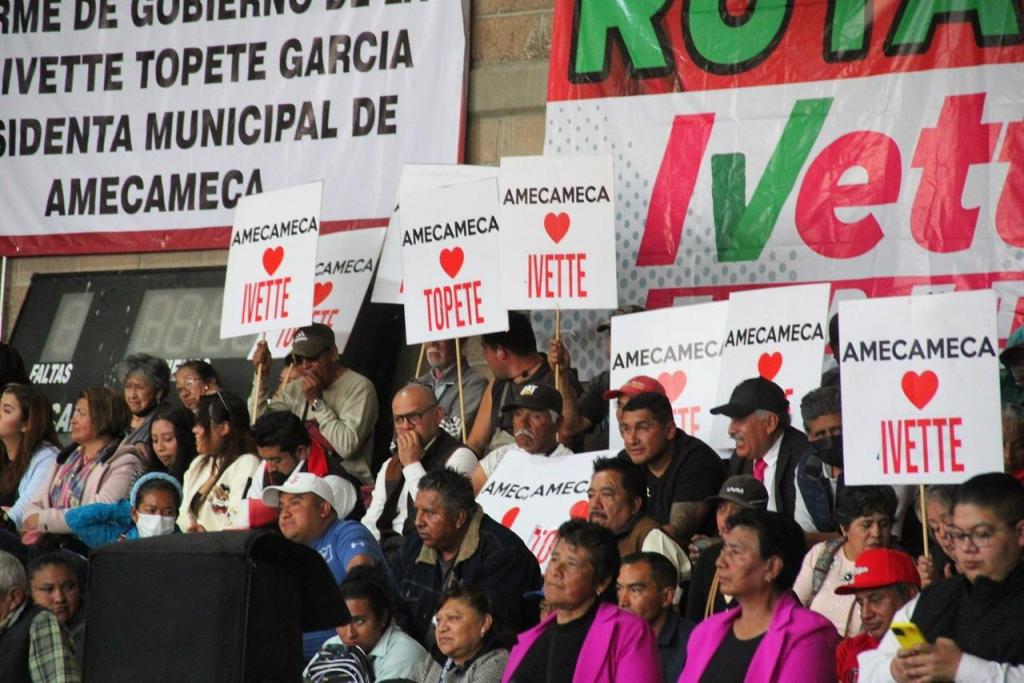 Un grupo de personas asiste a un evento, sosteniendo carteles que apoyan a la alcaldesa Ivette Topete García, con el mensaje 'Amecameca ♥ Ivette' y 'Amecameca ♥ Topete' de fondo.