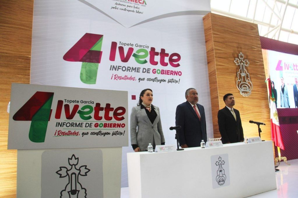 Evento de presentación del primer informe de Gobierno de la alcaldesa Ivette Topete García, con un fondo decorativo y la presencia de funcionarios, marcando los logros y avances en la administración municipal.