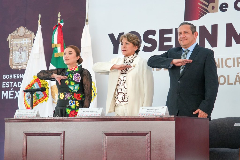 Tres personas de pie con la mano derecha levantada, en un evento formal, frente a un fondo con banderas y un logotipo de gobierno.