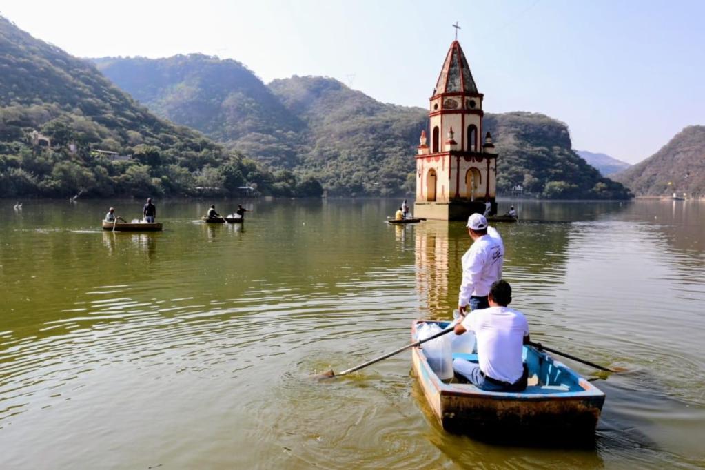 Grupo de personas en botes remando en un lago con montañas al fondo y una iglesia sumergida parcialmente en el agua.
