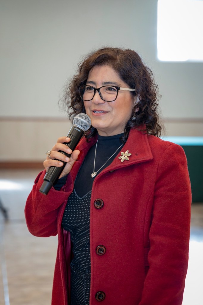 La rectora de la Universidad Autónoma del Estado de México habla frente a un micrófono, vestida con un abrigo rojo y blusa de puntos, en un contexto formal.