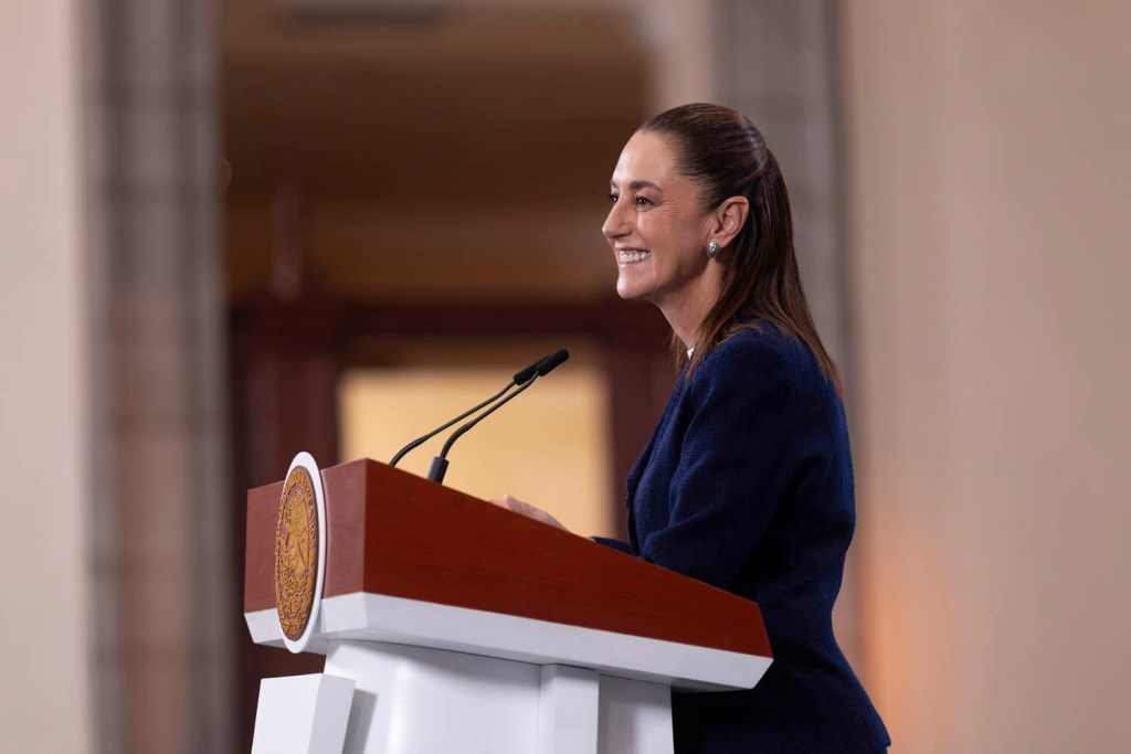 A woman standing at a podium, smiling and speaking, with a blurred background.