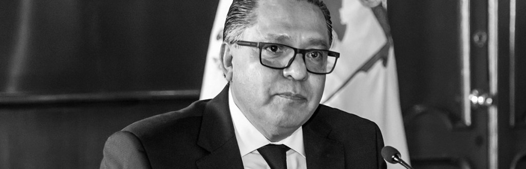 Retrato en blanco y negro de un hombre con gafas, sentado frente a un micrófono, con una expresión seria en un entorno formal.