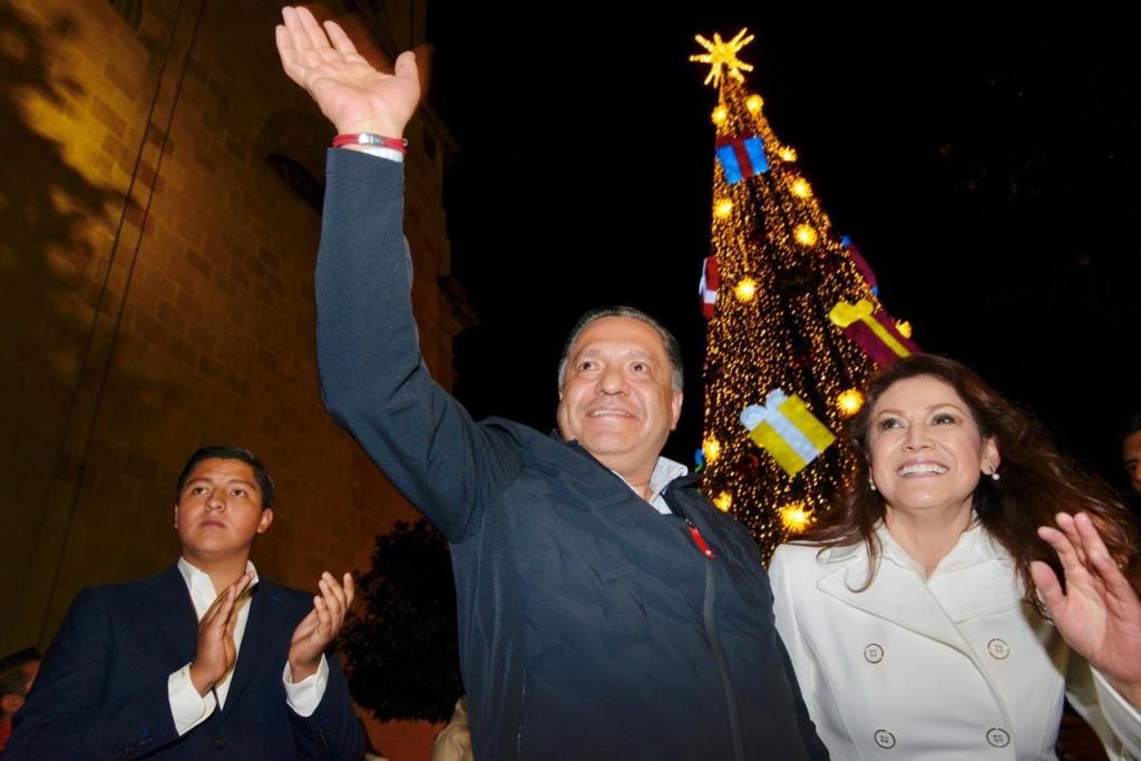 El presidente municipal Ricardo Moreno y la presidenta del DIF Toluca Rocío Pegueros Velázquez saludan al público durante el encendido del Árbol Navideño en Toluca, con luces brillantes y regalos decorativos en el fondo.