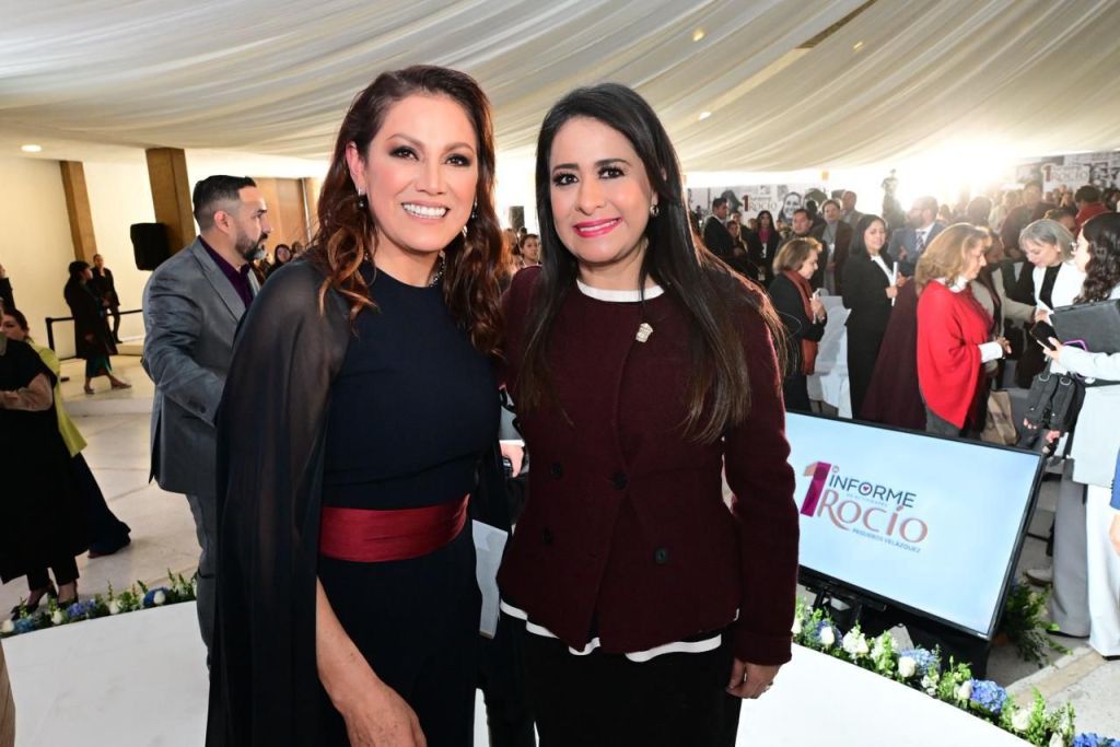 Rocío Pegueros Velázquez y otra mujer sonriendo en el evento de presentación del Primer Informe de Actividades del DIF Toluca, rodeadas de asistentes y decoraciones festivas.