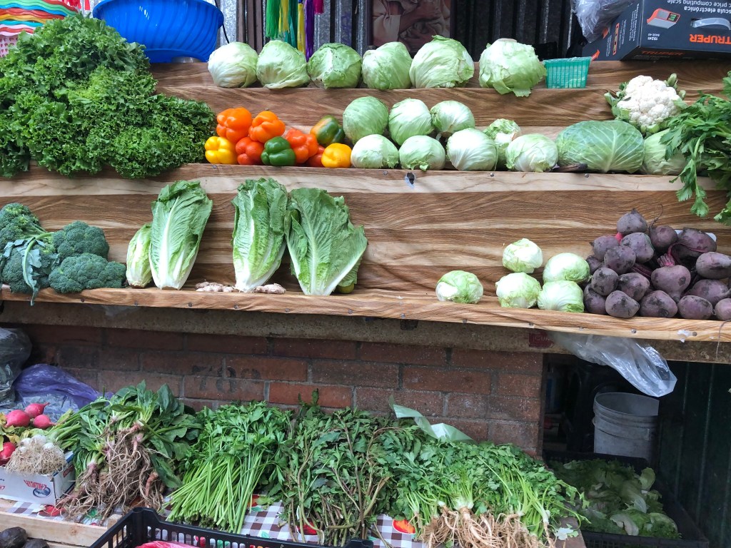 Un mercado colorido con una variedad de verduras frescas, incluyendo lechuga, brócoli, col, cebollas, y betabel, exhibidas en estantes de madera en un ambiente urbano.