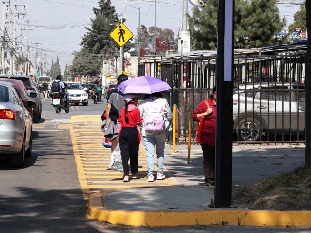 Dos niñas caminando por la calle, una de ellas sostiene un paraguas morado mientras pasan junto a un coche en el lado izquierdo. Se observan otros peatones y tráfico en la carretera.
