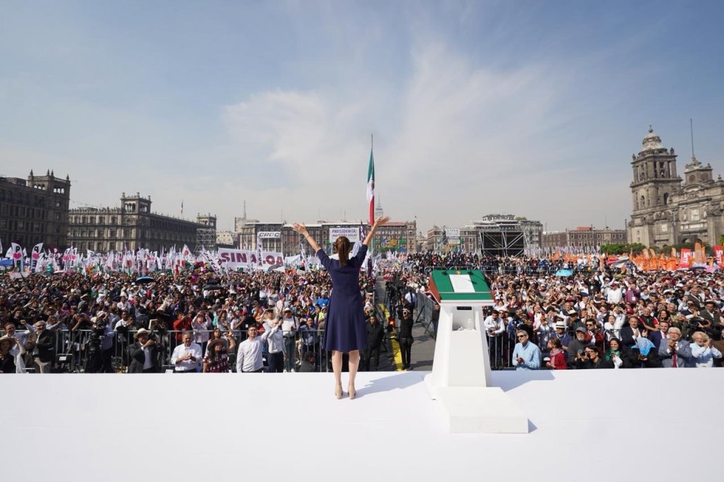 La presidenta Claudia Sheinbaum encabezando un mitin en el Zócalo de la Ciudad de México, con una gran multitudes de personas y banderas ondeando.