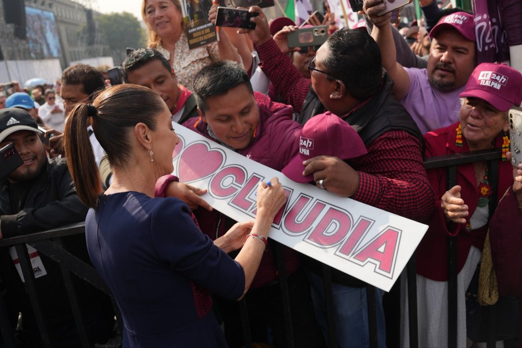 Claudia Sheinbaum firmando un cartel con 'I ❤️ CLAUDIA' mientras interactúa con sus seguidores en un evento político, rodeada de personas que la apoyan.