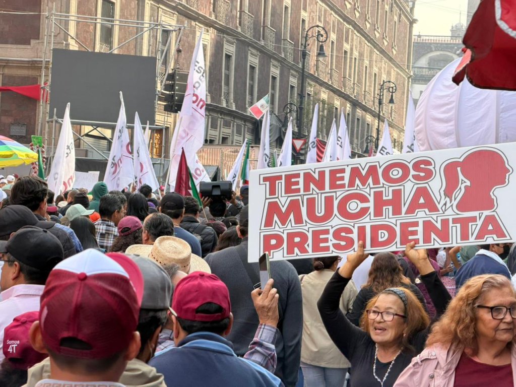 Multitudinaria manifestación en el Zócalo de la Ciudad de México, con personas sosteniendo un cartel que dice 'TENEMOS MUCHA PRESIDENTA'. Se observan banderas y un ambiente de celebración política.