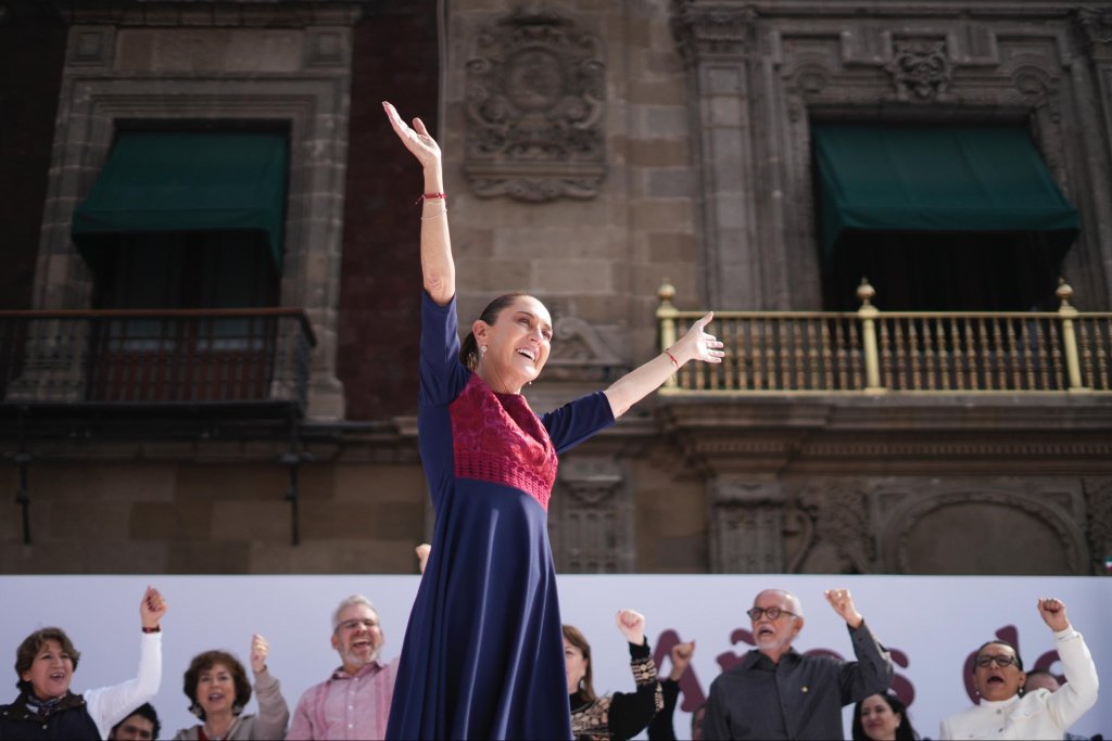 Claudia Sheinbaum se encuentra en el Zócalo, levantando los brazos con entusiasmo durante su discurso en conmemoración de los siete años del triunfo de López Obrador, rodeada de un grupo de personas que la apoyan.