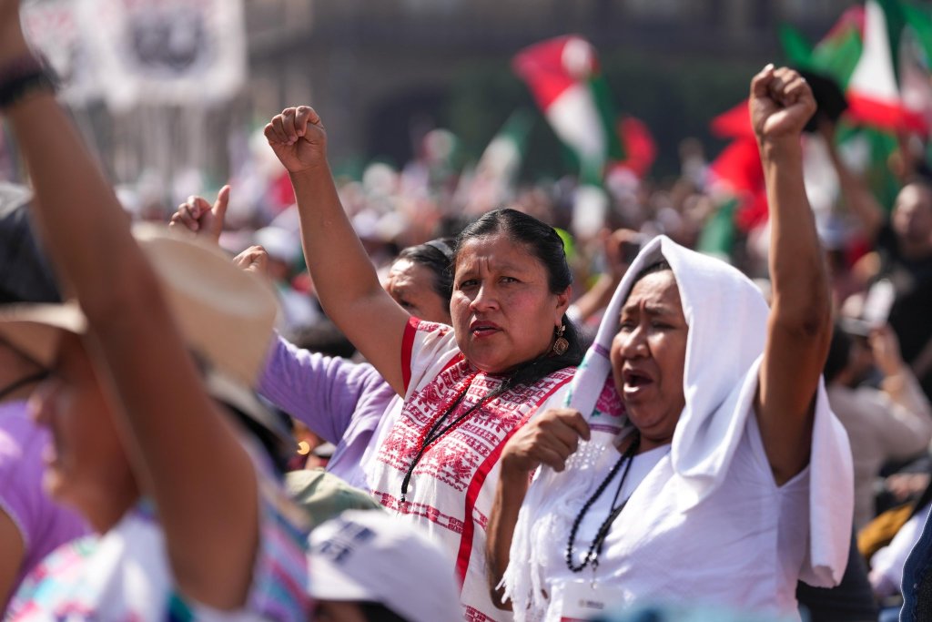 Grupo de mujeres levantando los puños en señal de protesta o apoyo, vestidas con ropa tradicional, en un evento multitudinario al aire libre.