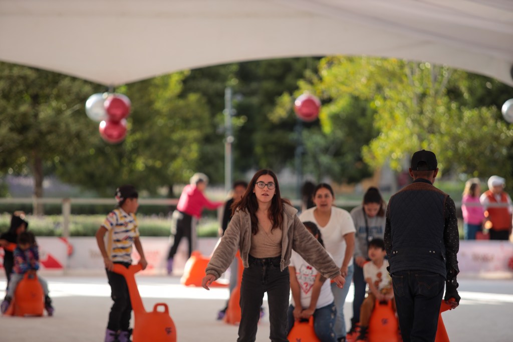 Grupo de personas patinando sobre hielo en una pista al aire libre, con bolos de entrenamiento y decoraciones navideñas en el fondo.
