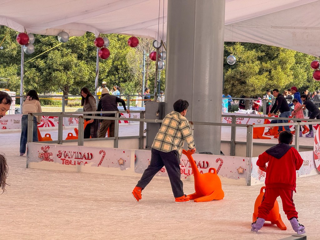 Patinadores disfrutando de una pista de hielo en Toluca, con decoraciones navideñas y asistencia de sopor para los novatos.