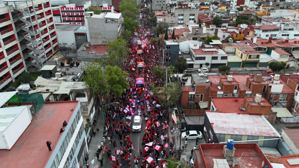 Vista aérea de una multitud de personas en una celebración, con banderas y pancartas, en una calle rodeada de edificios y casas.