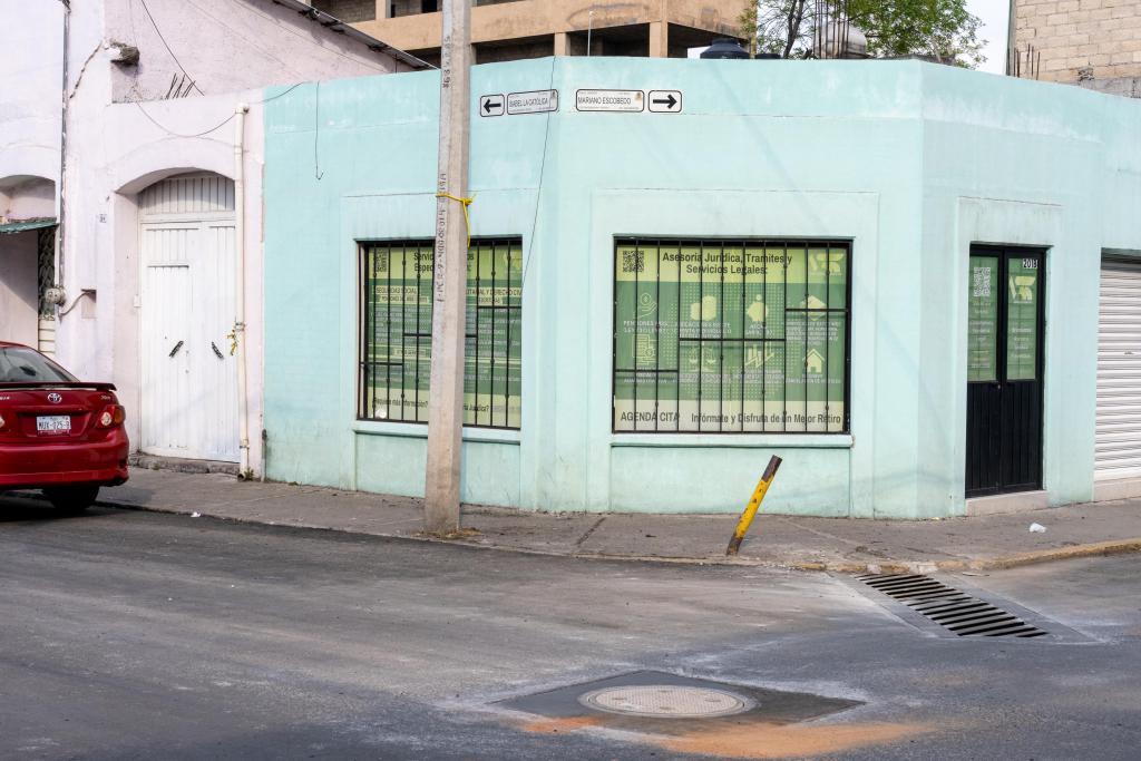 Esquina de un edificio color azul claro con ventanas enrejadas, ubicado en una calle, con un automóvil rojo estacionado frente a la entrada.