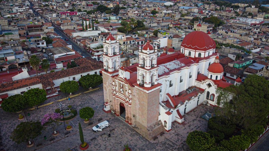 Vista aérea de una iglesia con cúpulas rojas y arquitectura colonial, rodeada de un paisaje urbano en el sur del Estado de México.