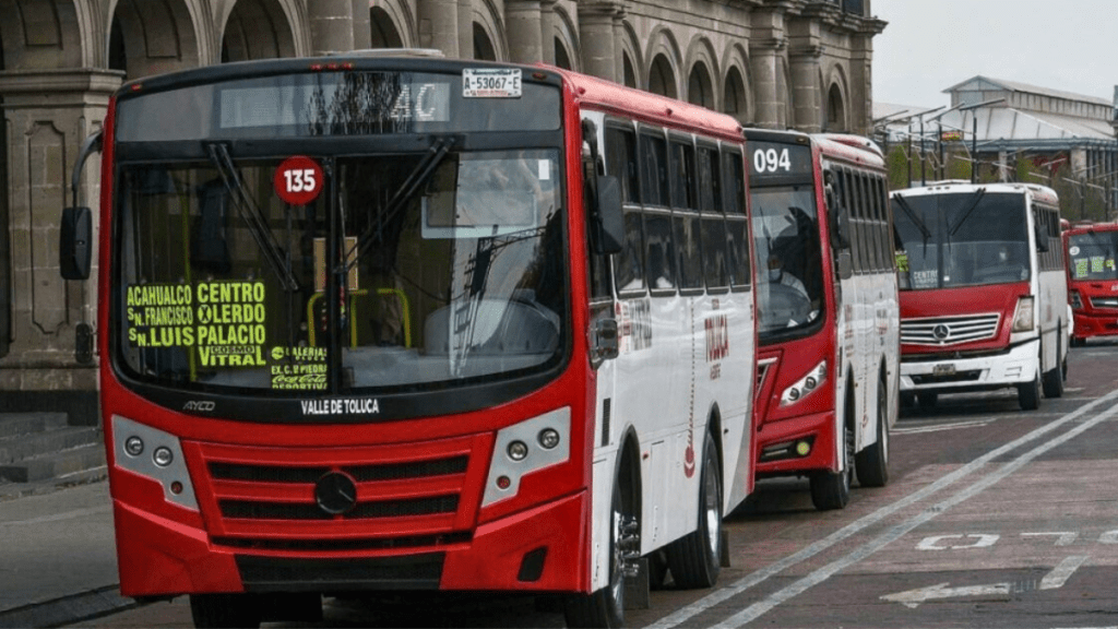 A line of red and white public buses parked on a street in a city, displaying route numbers and destinations on their front panels.