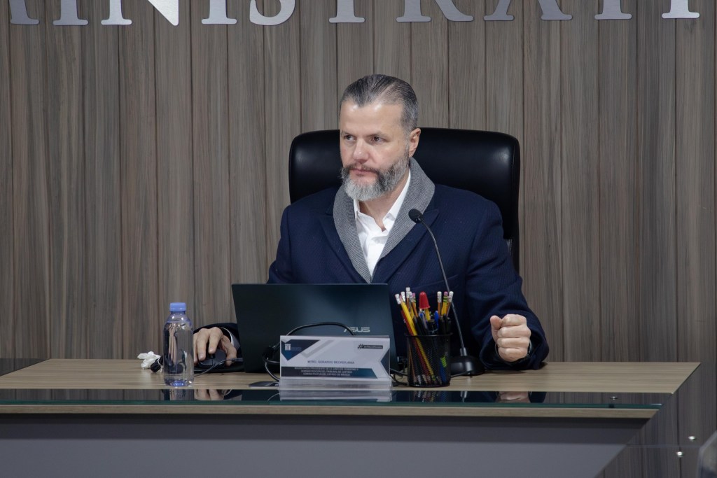 Retrato de un hombre sentado en un escritorio, con una computadora portátil y utensilios de oficina, en un ambiente de reunión formal.