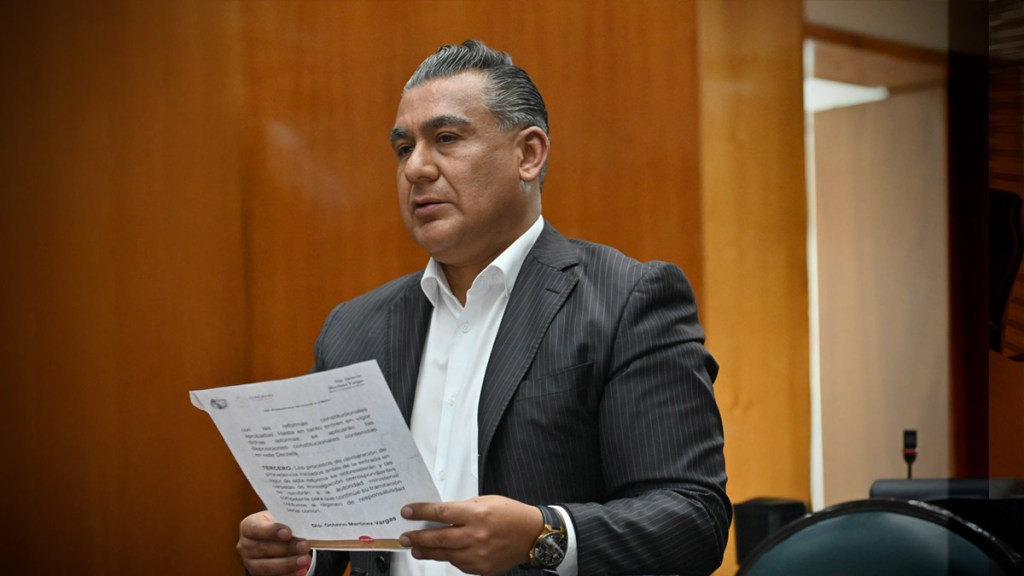 Un hombre sosteniendo un documento frente a un fondo de madera, posiblemente en un entorno de trabajo o reunión.