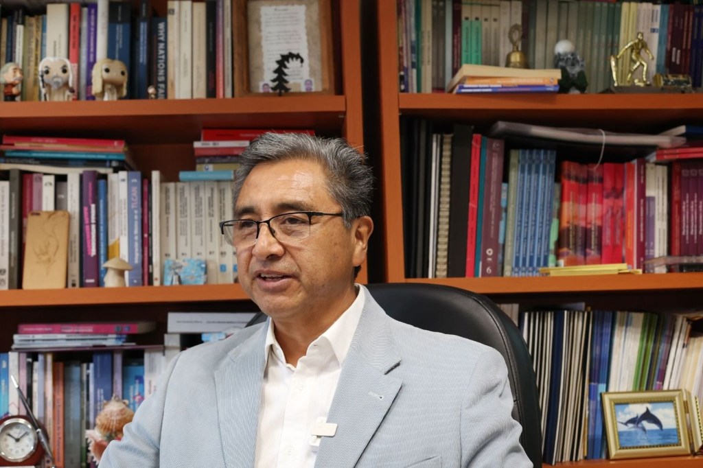 Retrato de Pablo Mejía Reyes, coordinador del Centro de Investigación en Ciencias Económicas, en su oficina con estanterías llenas de libros y objetos decorativos.