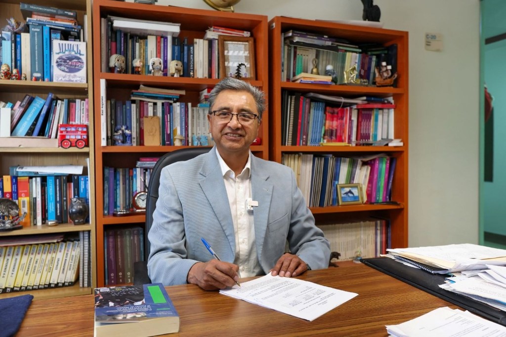 Un académico sentado en un escritorio, rodeado de estanterías repletas de libros y documentos, sosteniendo un bolígrafo y mirando a la cámara con una sonrisa.
