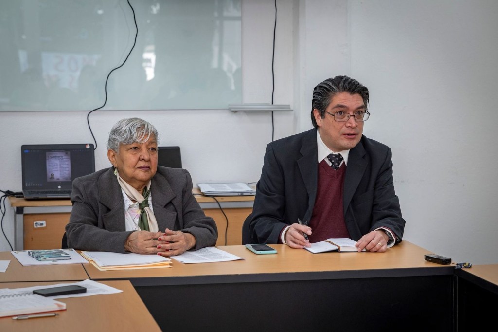 Dos personas sentadas frente a una mesa, una mujer mayor con cabello blanco y un hombre de mediana edad con gafas, ambos escuchando atentamente en un entorno académico.