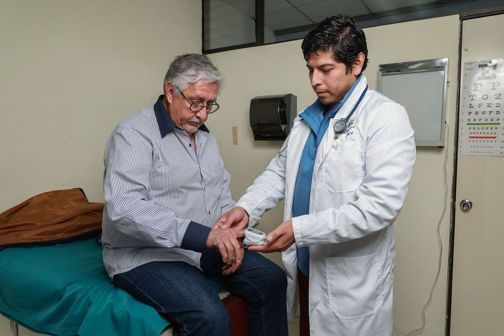 Un médico en bata blanca está revisando a un paciente mayor sentado en una cama de consulta médica.