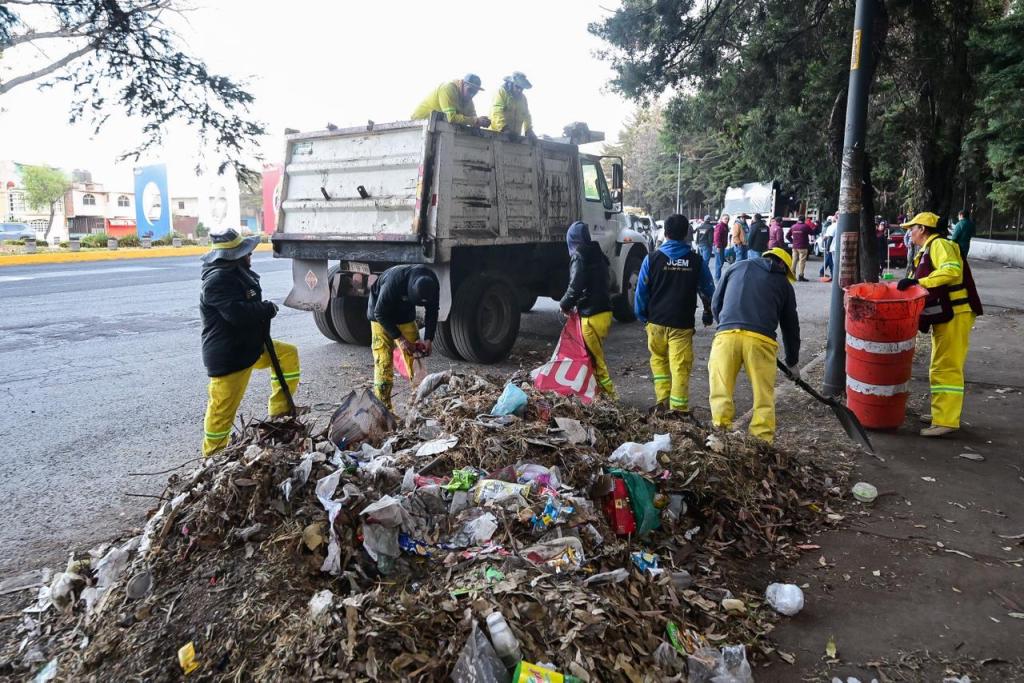 Grupo de trabajadores de limpieza en ropa amarilla recogiendo basura en la calle, con un camión de recolección de residuos en el fondo.