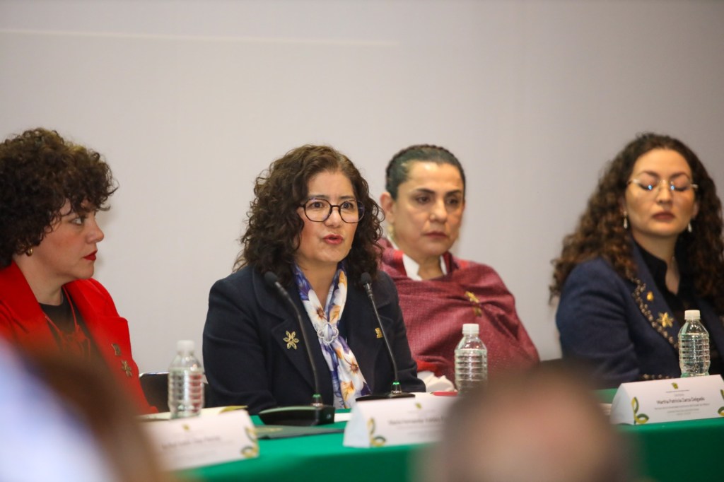 A panel of four women at a conference table, with one woman speaking into a microphone and the others listening attentively.