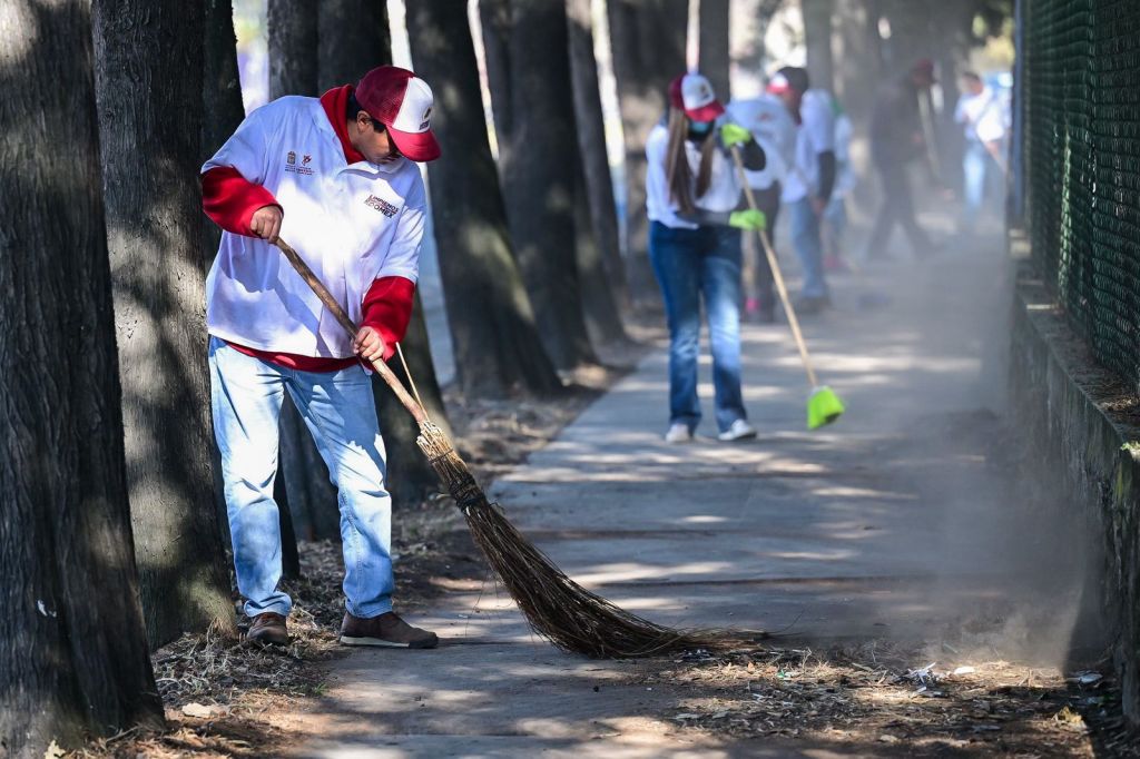 Un grupo de personas limpiando una acera, barrendo hojas y escombros, en un ambiente al aire libre. Se ven árboles a los lados y algunos voluntarios usando camisetas blancas y gorras.