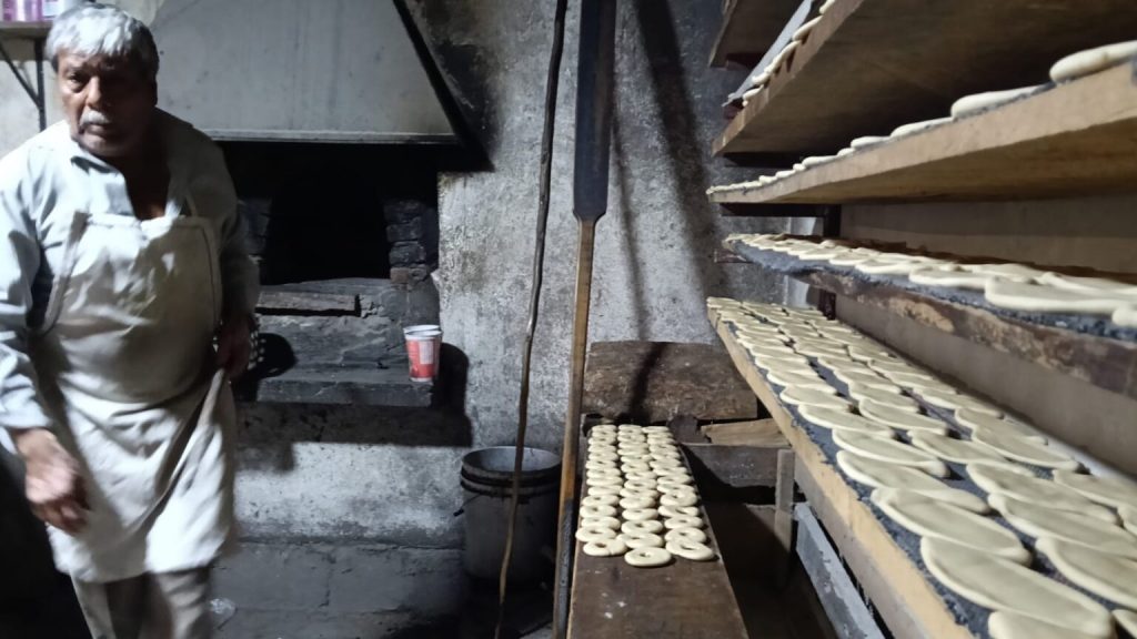 Hombre mayor preparando cocoles en un horno de piedra, con piezas de masa listas en estantes de madera.