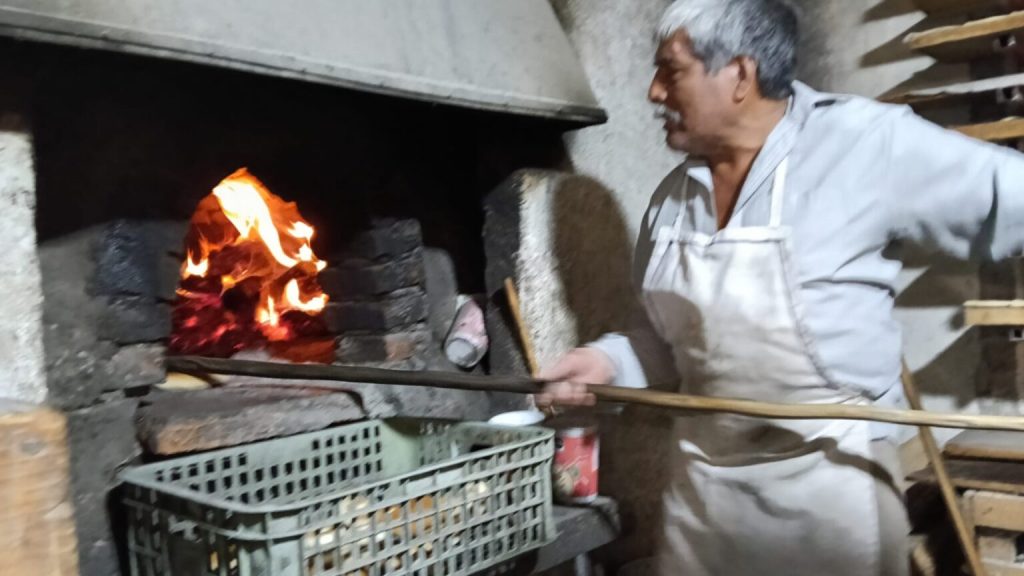 Un panadero frente a un horno de piedra ardiendo, sosteniendo una herramienta de madera, mientras supervisa el proceso de horneado.