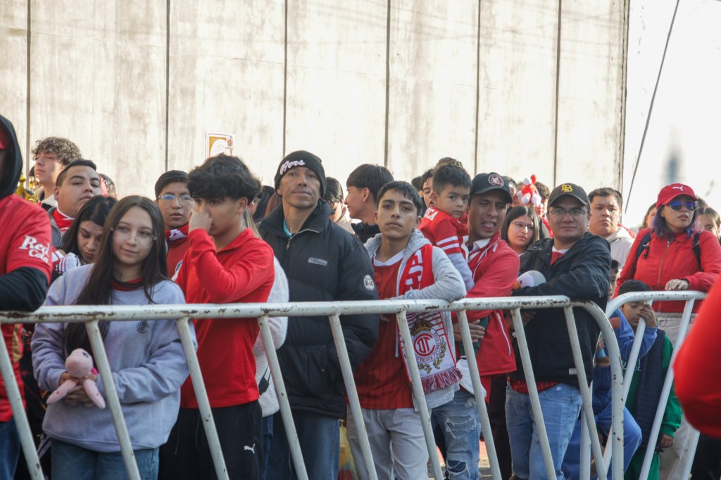 Grupo de aficionados esperando en la entrada del Estadio Nemesio Diez, algunos con ropa del Toluca FC y otros sosteniendo juguetes para donar.