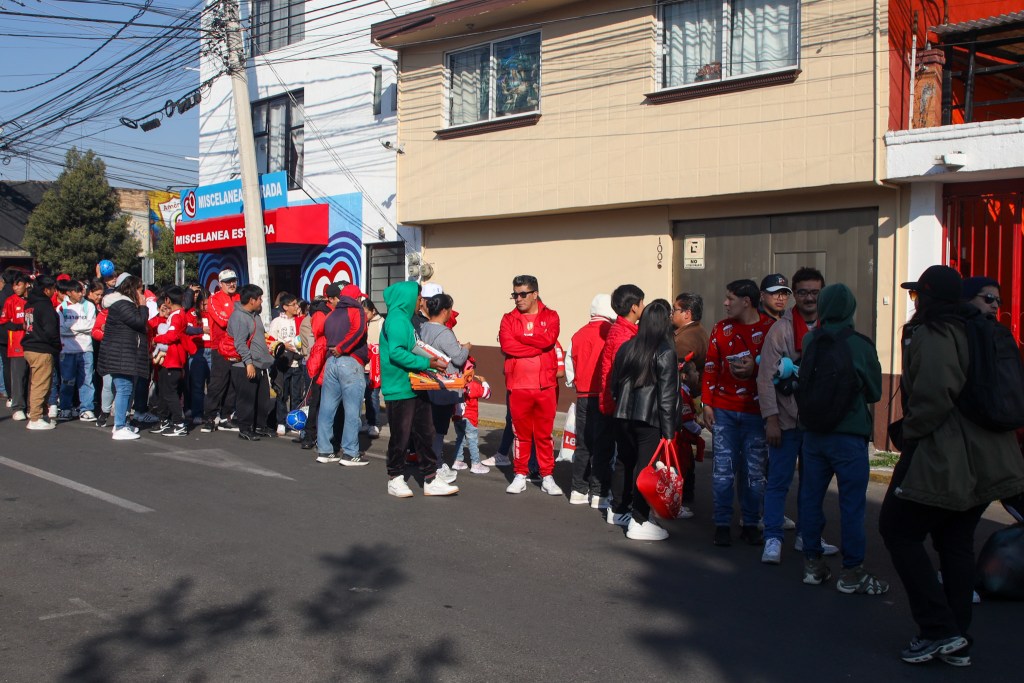 Una larga fila de aficionados del Toluca FC esperando para entrar al estadio, muchos de ellos vestidos con camisetas rojas y llevando juguetes para donar.