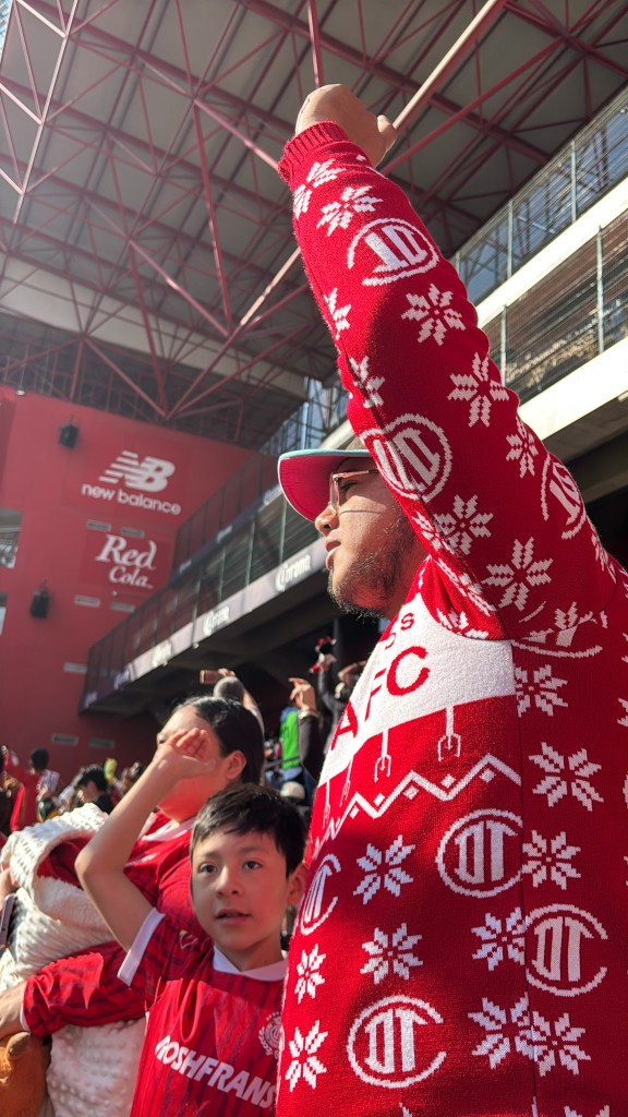 Un aficionado del Toluca FC vestido con un suéter navideño rojo levanta el brazo con entusiasmo mientras un niño lo mira con alegría, todo en un ambiente festivo en el estadio.