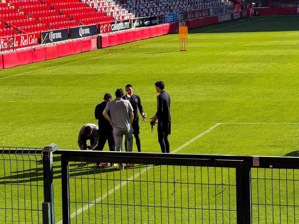 Entrenamiento en el Estadio Nemesio Diez con varios jugadores discutiendo en el campo, rodeados de un césped verde y asientos rojos.