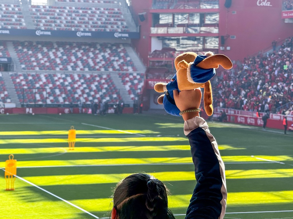 Una persona en el estadio levanta un peluche en un ambiente festivo mientras se lleva a cabo un entrenamiento del Toluca FC.