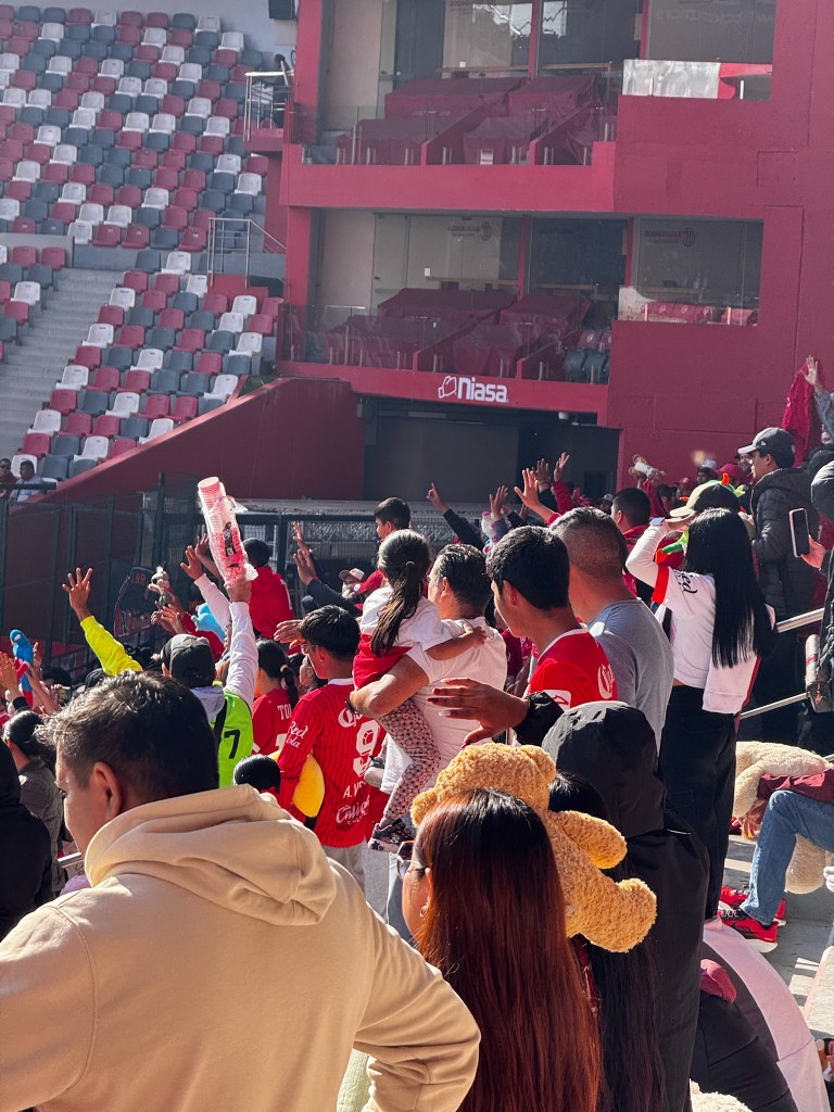Aficionados del Toluca FC animan desde las gradas durante un entrenamiento especial en el Estadio Nemesio Diez, vistiendo ropa en colores del equipo y sosteniendo peluches para donar.