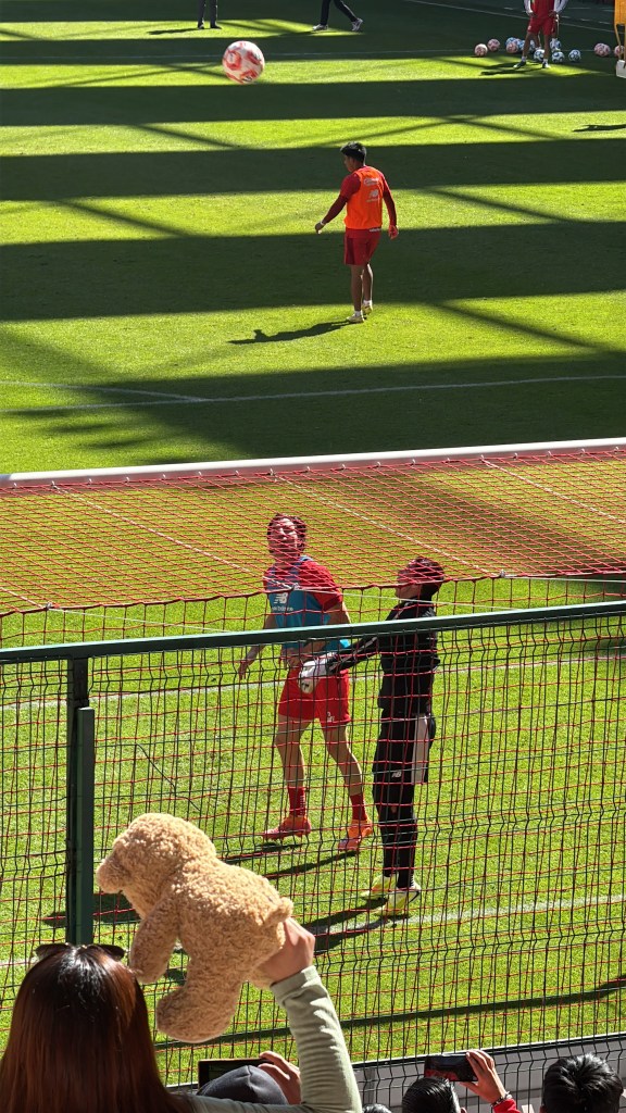 Jugador del Toluca FC entrenando mientras un aficionado sostiene un peluche en la tribuna.