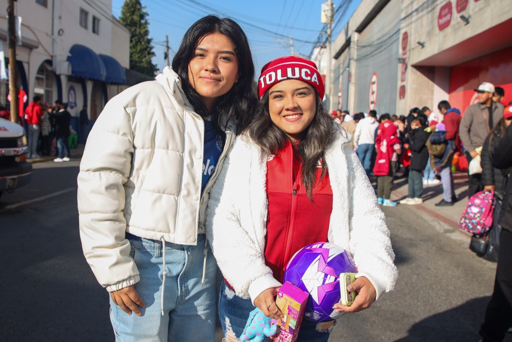 Dos jóvenes sonrientes posando juntas en la calle, vistiendo prendas de abrigo. Una de ellas lleva un gorro rojo con la palabra 'Toluca', mientras sostiene un balón de fútbol de colores y una caja de donación. Al fondo, se observan personas en fila para participar en el evento.