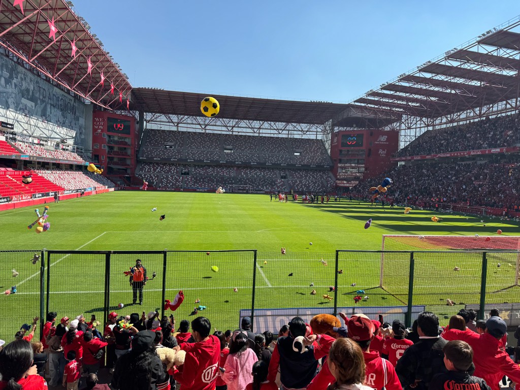 Vista del Estadio Nemesio Diez lleno de aficionados en un evento de entrenamiento, con peluches lanzados al campo como parte de una colecta para donar a niños, bajo un cielo despejado.