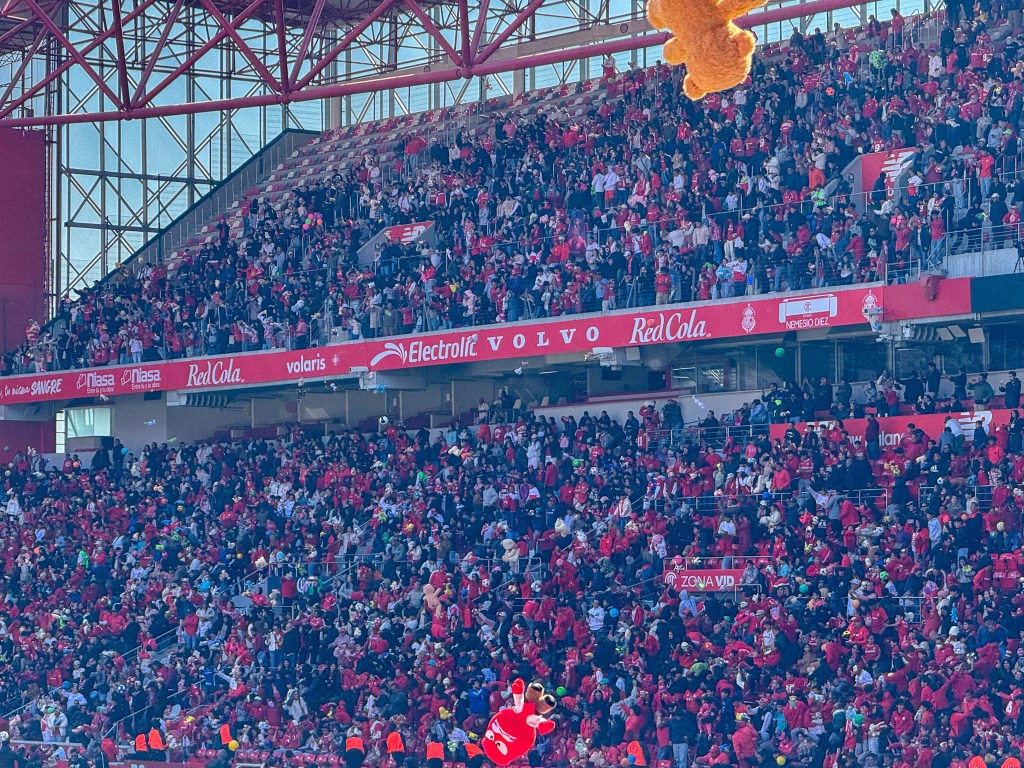 Aficionados del Toluca FC en el Estadio Nemesio Diez durante un evento especial, con una multitud de personas vestidas de rojo y un peluche volando en el aire.