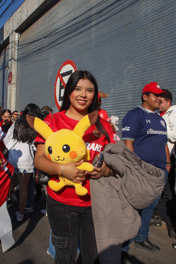 Una aficionada del Toluca FC sonríe mientras sostiene un peluche de Pikachu, vistiendo una camiseta roja del equipo, en un evento de recolección de juguetes.