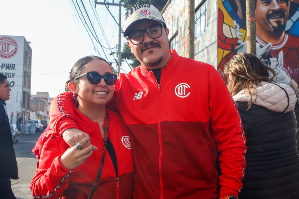 Pareja sonriente con ropa del Toluca FC, posando en un ambiente festivo con murales en el fondo.