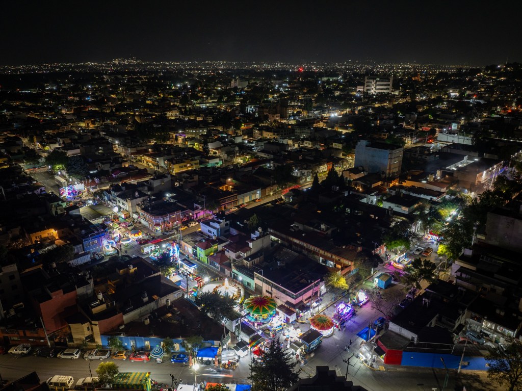 Vista aérea de una ciudad iluminada por la noche, con calles llenas de luces y un ambiente festivo. Se pueden ver ferias y atracciones en la parte inferior de la imagen.