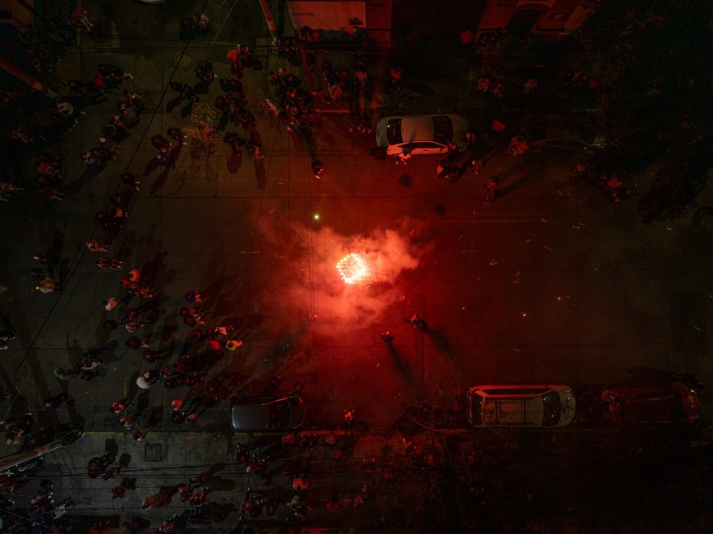 Vista aérea de una calle con gente reunida y fuegos artificiales arrojando humo rojo.