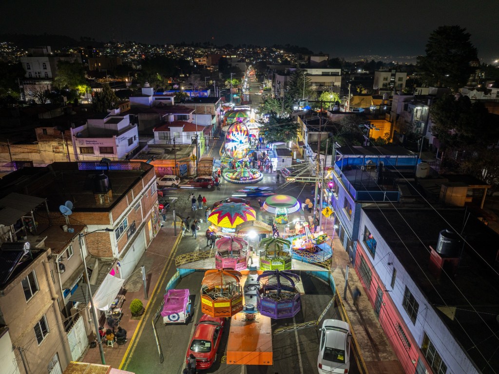 Vista nocturna de una calle iluminada con luces coloridas y decoraciones festivas, donde se pueden ver ferias y atracciones, y personas paseando en un ambiente alegre.