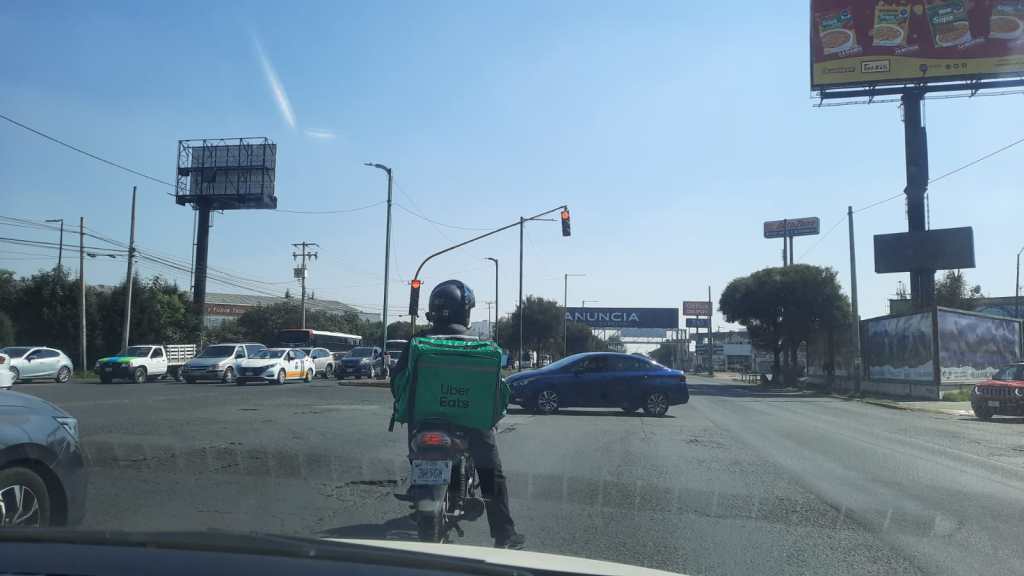 Motociclista con mochila de Uber Eats esperando en un semáforo, con vehículos en movimiento y un panorama urbano en Toluca.